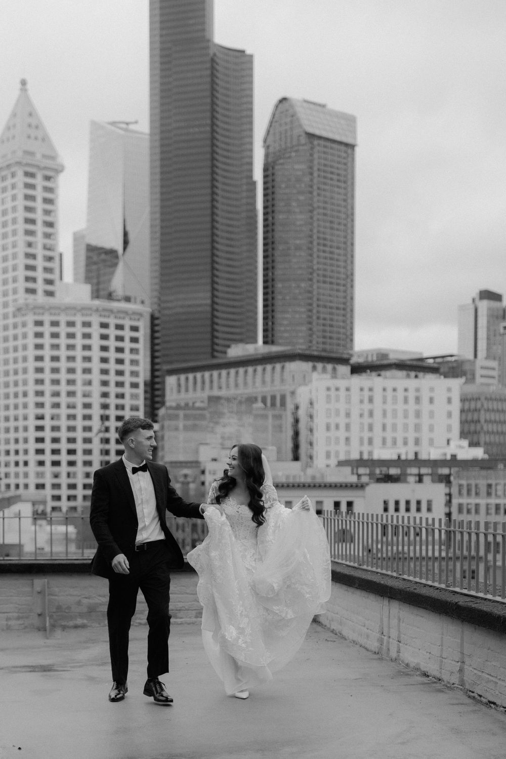 A couple in wedding attire walks together on a rooftop with the Seattle skyline in the background, black and white photo.