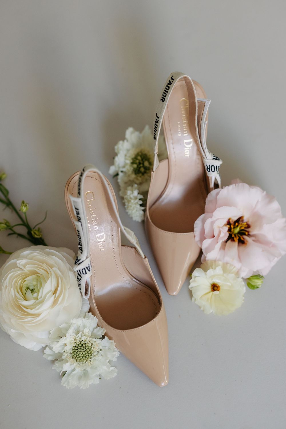 Elegant nude Dior slingback heels styled with delicate white and pink flowers on a light gray background.