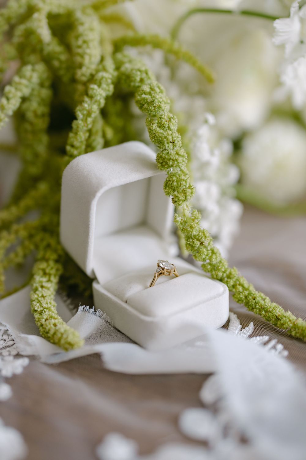 Diamond engagement ring in white velvet box with green amaranthus flowers and white decor details.
