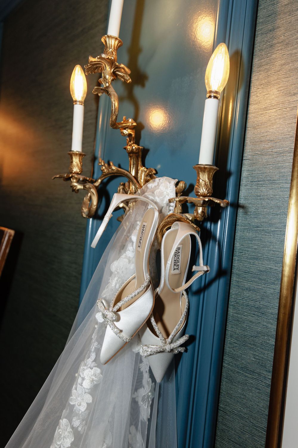 A white lace wedding dress and bridal heels hang elegantly against a blue wall with ornate gold candlestick sconces.