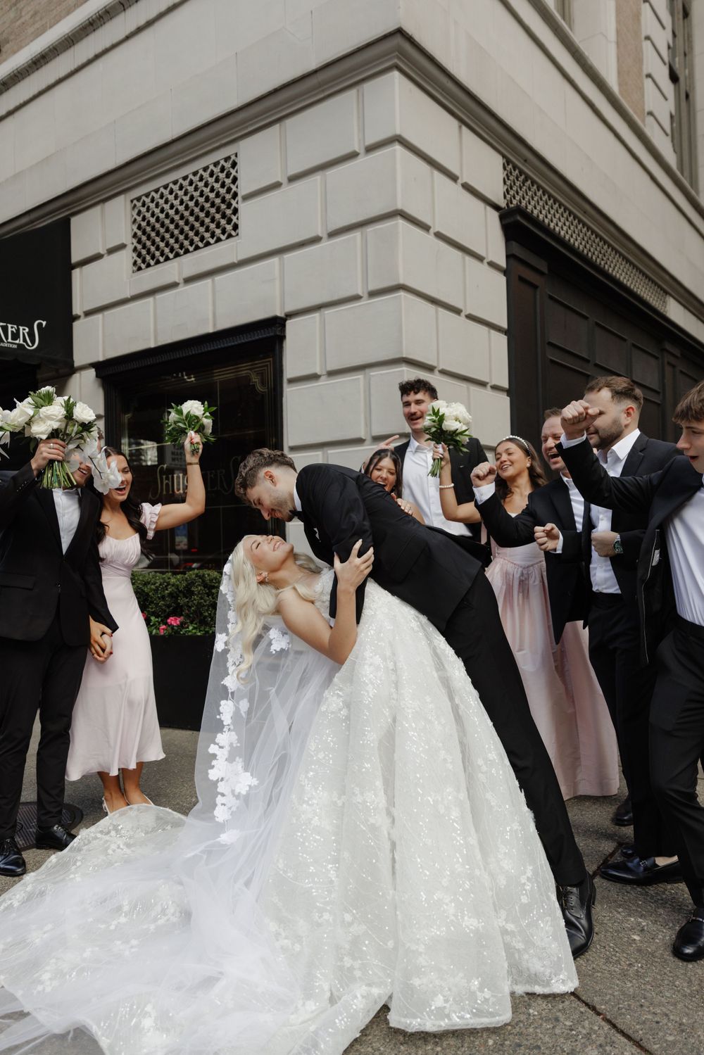 Wedding party celebrates outside building as couple shares romantic dip kiss in elegant white gown and formal attire.