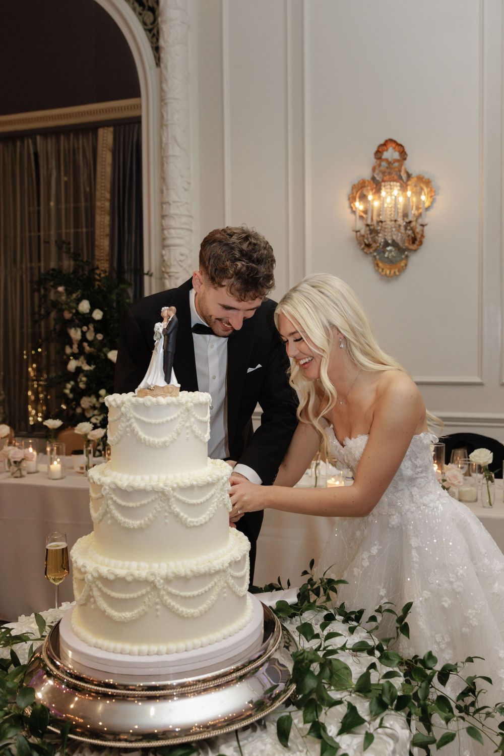 Newlyweds prepare to cut their elegant four-tier white wedding cake decorated with floral and pearl details on a silver stand.