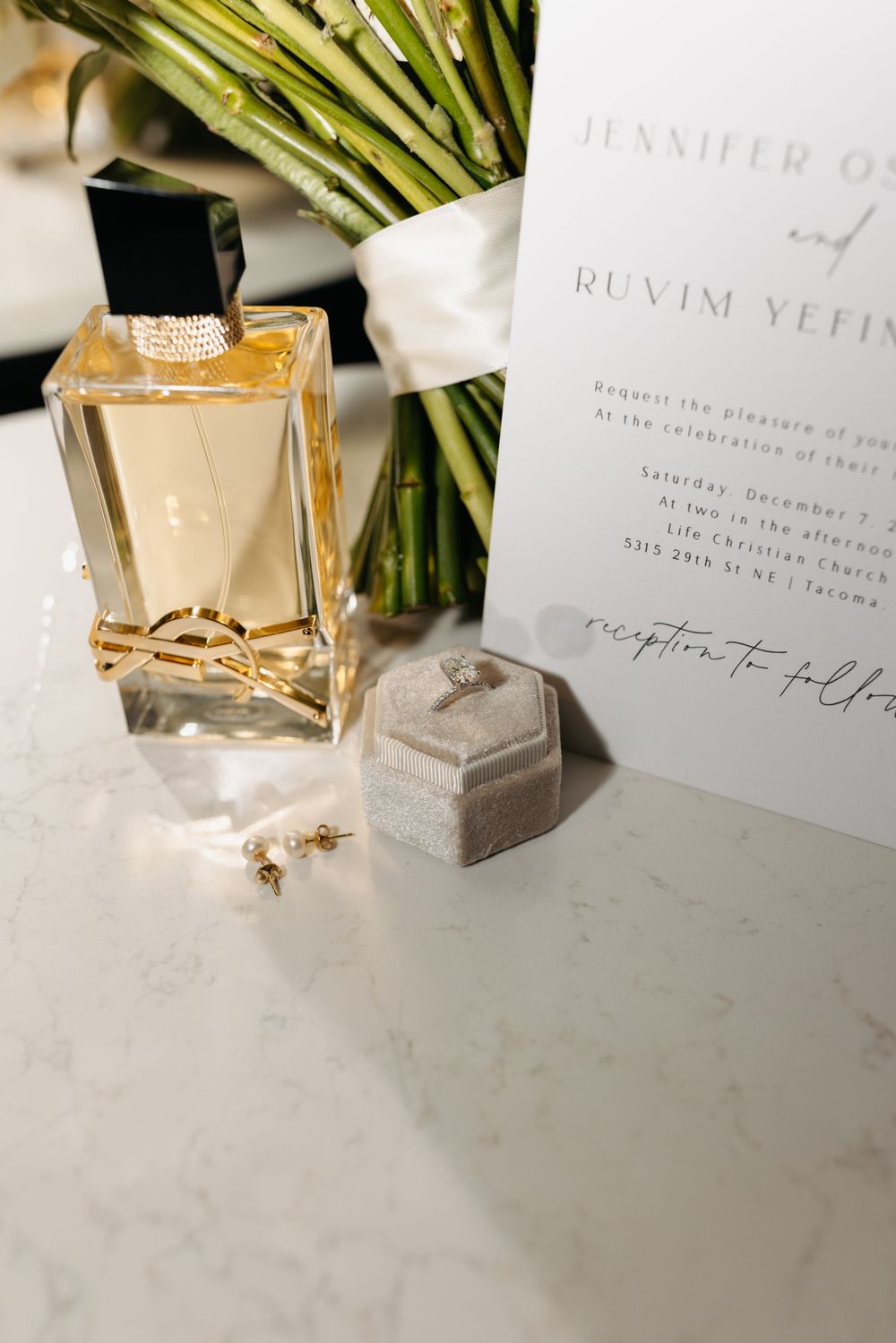Wedding details including a perfume bottle, ring box, invitation and greenery arranged on white marble surface.