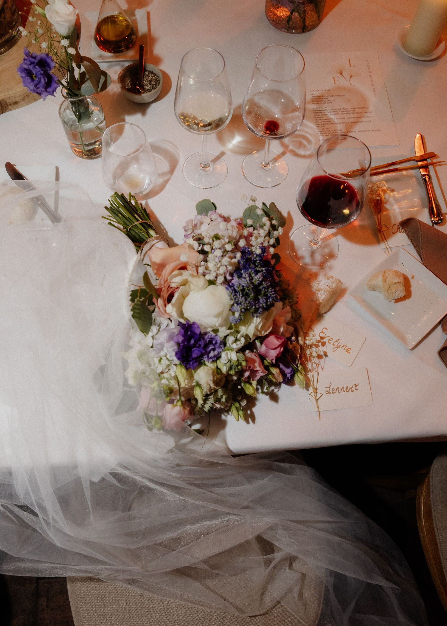 Elegante place setting met wijnglazen en verspreide witte en paarse bloemstukken gedrapeerd in vloeiende pure stof.