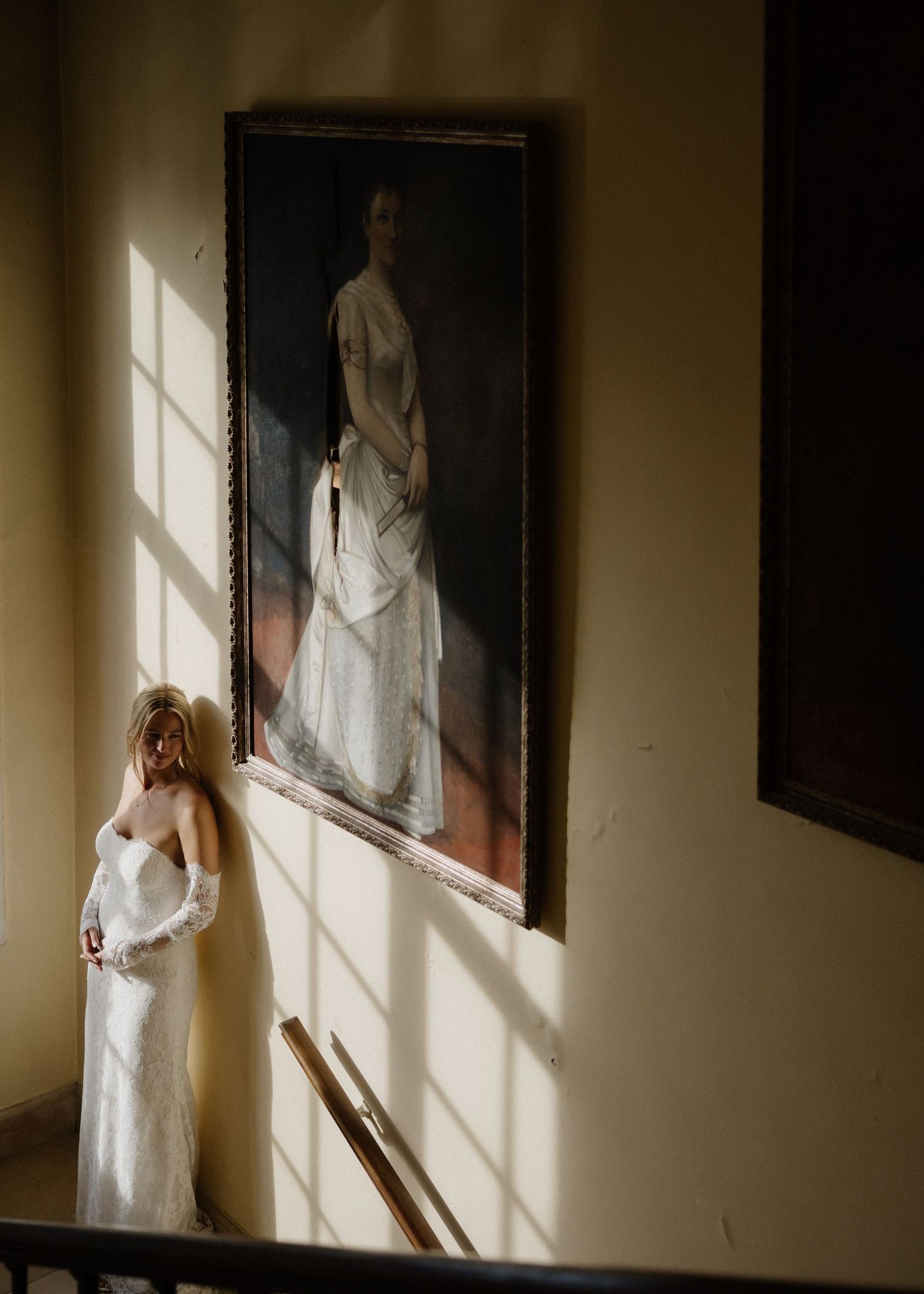 Bride in white gown stands beside a large portrait painting, bathed in dramatic sunlight inside an elegant venue.