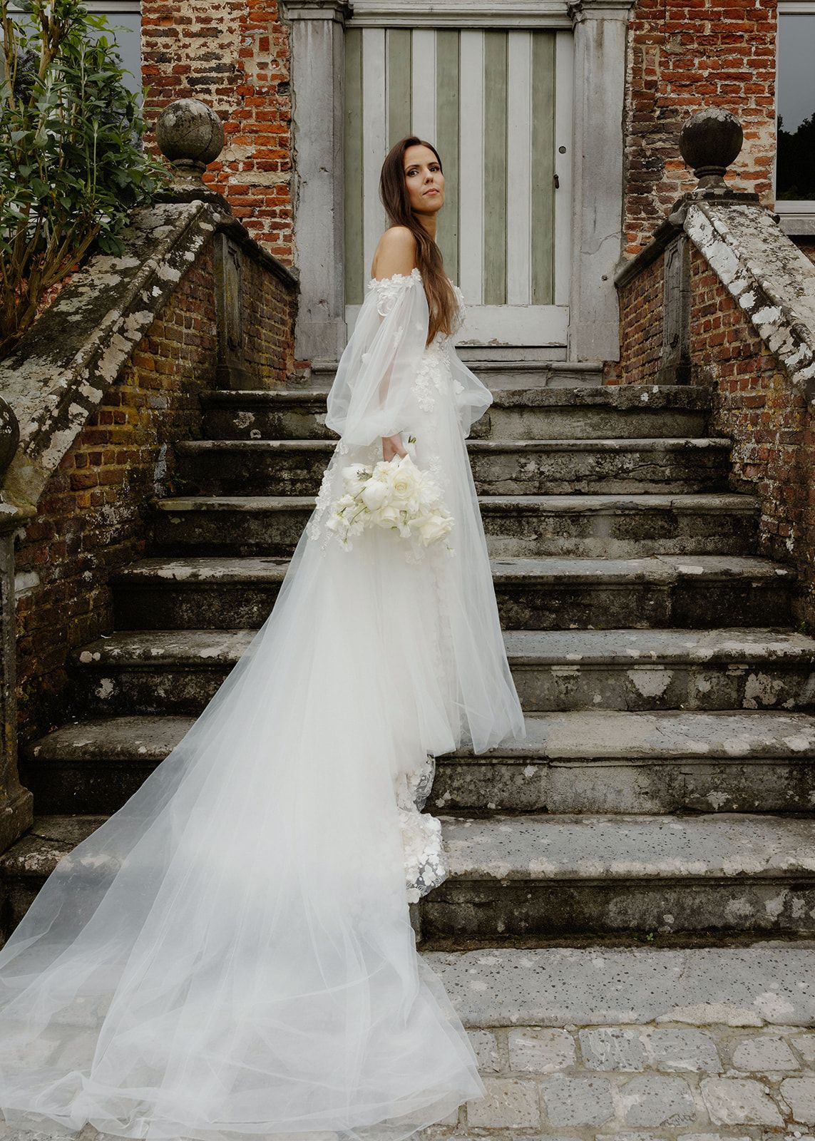 Bruid in off-shoulder jurk met lange sleep poseert op historische stenen trappen met wit boeket in Abbeye de Nizelles.