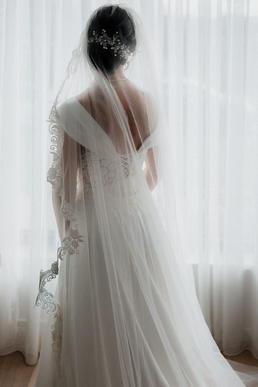 Elegante  bruid in witte trouwjurk met kanten sluier poseert in de buurt van pure gordijnen in dromerig silhouet met achtergrondverlichting.