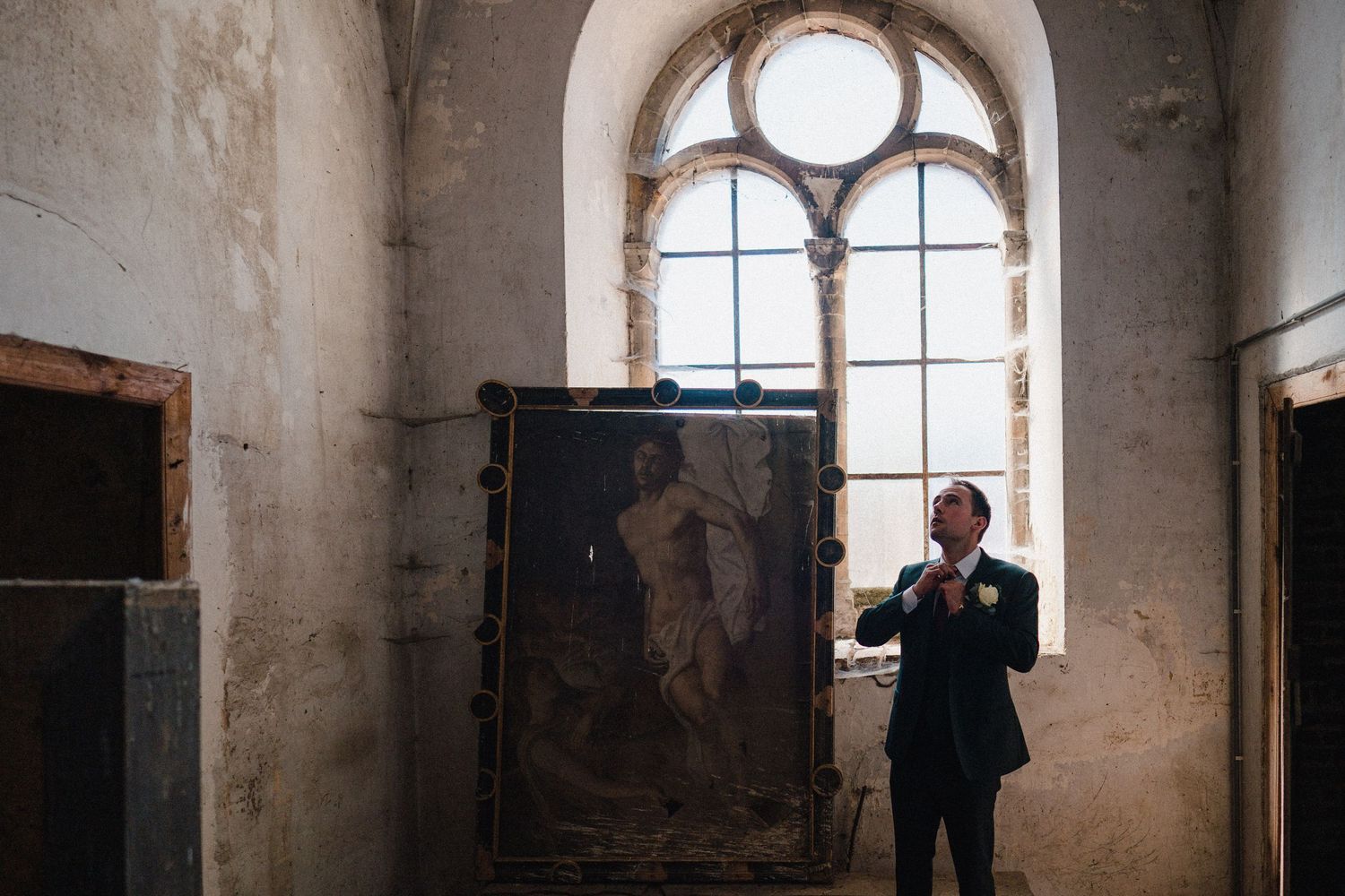 Man in suit adjusting tie beside large classical painting in ornate abandoned building with arched window.