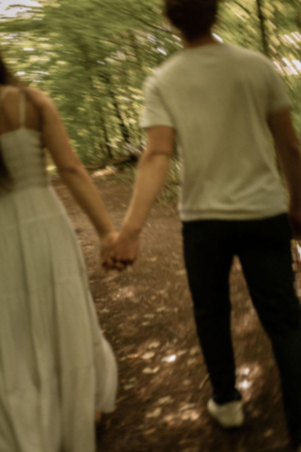 A couple holds hands while walking through a lush forest path, the woman in a white dress and man in casual attire.