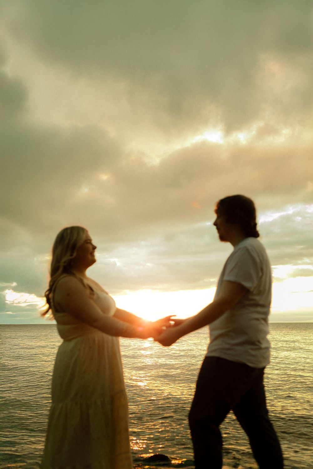 Pregnant couple holding hands at ocean sunset, silhouette against dramatic cloudy sky.