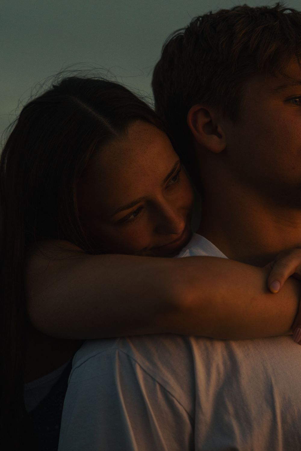A young couple shares a tender embrace at dusk, the woman resting her head on the man's shoulder.