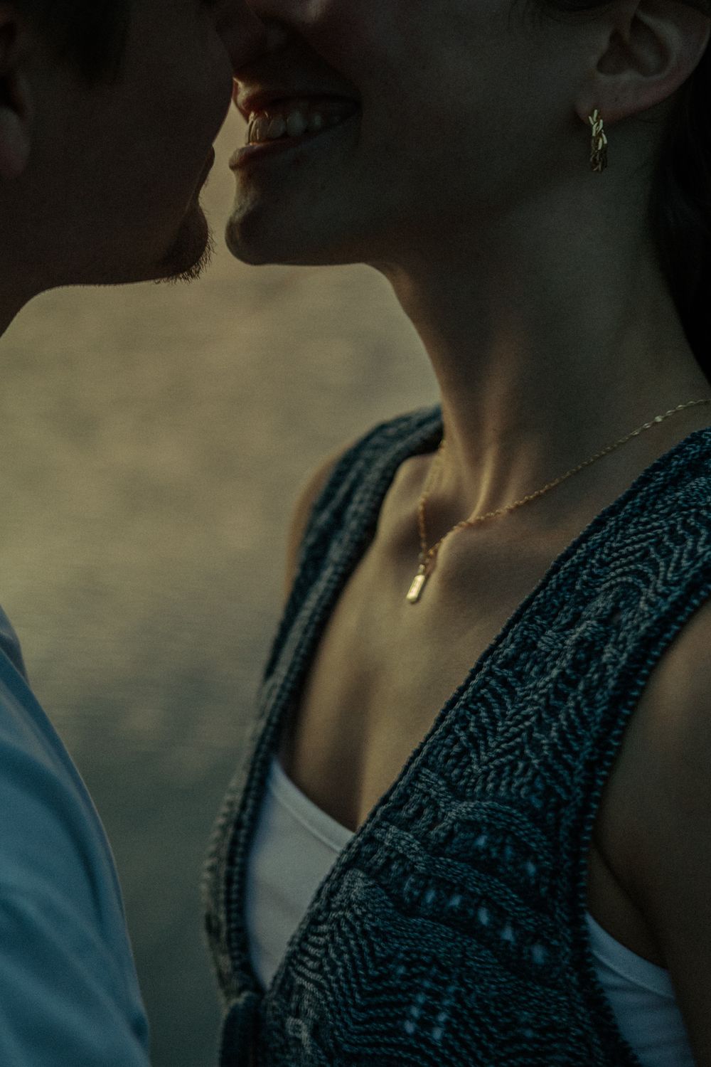 Close-up of a couple almost kissing, woman wearing a gold pendant necklace and earring in a teal knit vest.