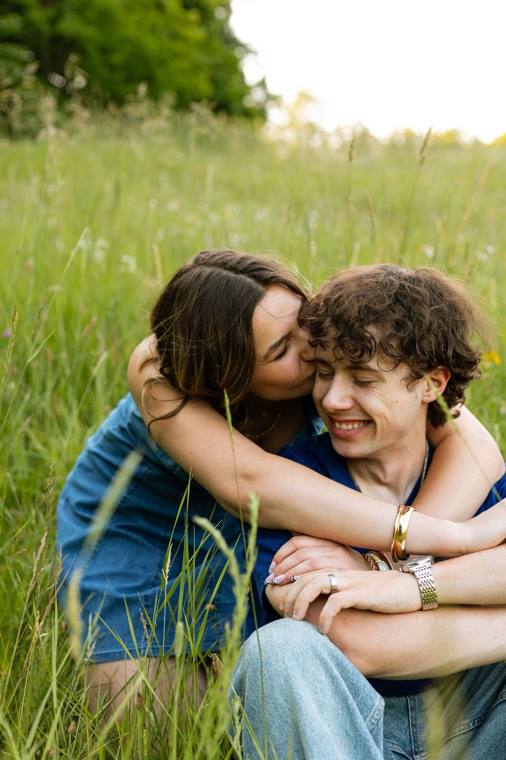 A couple sitting in a green meadow, woman kissing man on cheek while embracing him from behind.