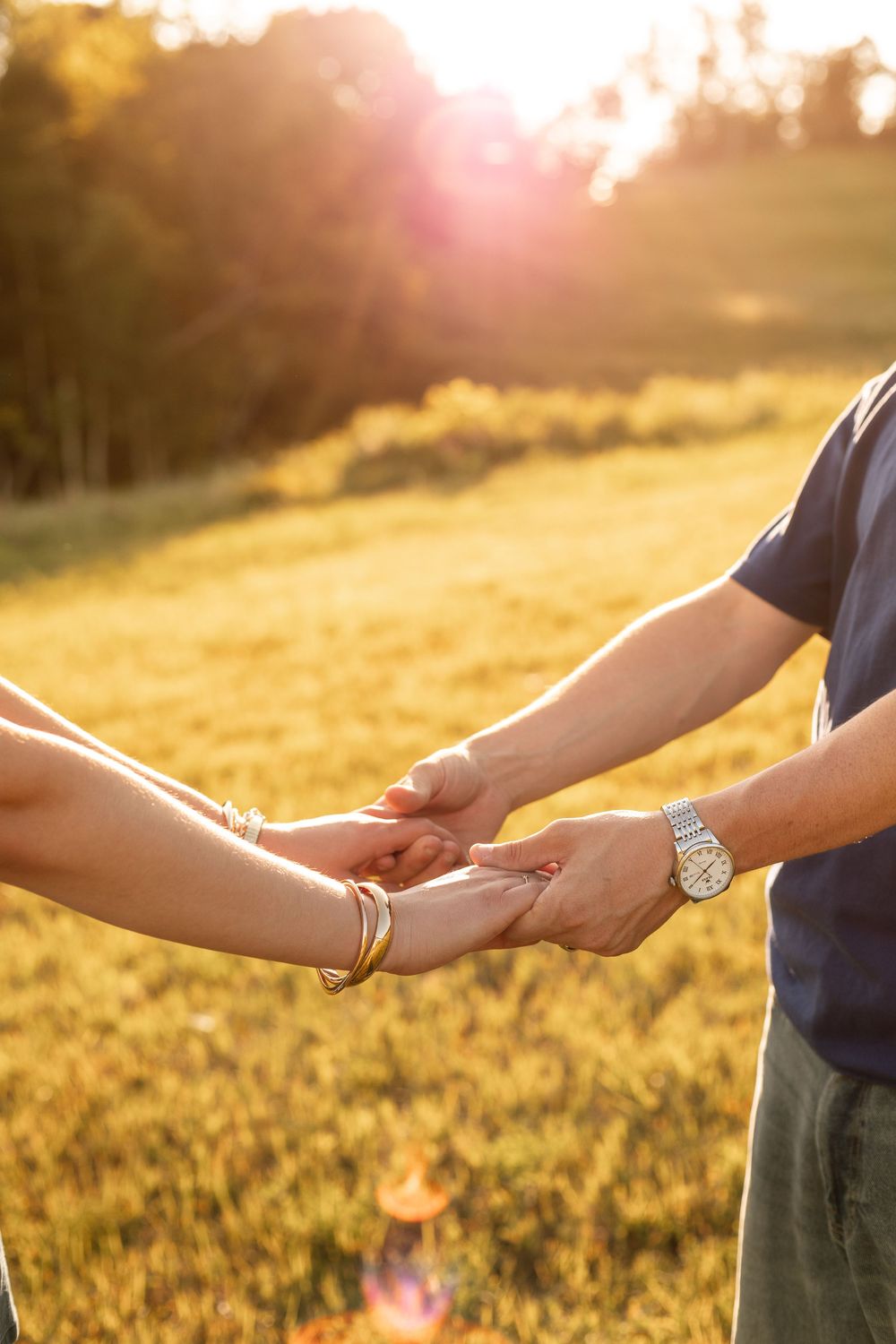 Couple holding hands tenderly in a golden sunset field, woman wearing gold bracelet, man with silver watch.