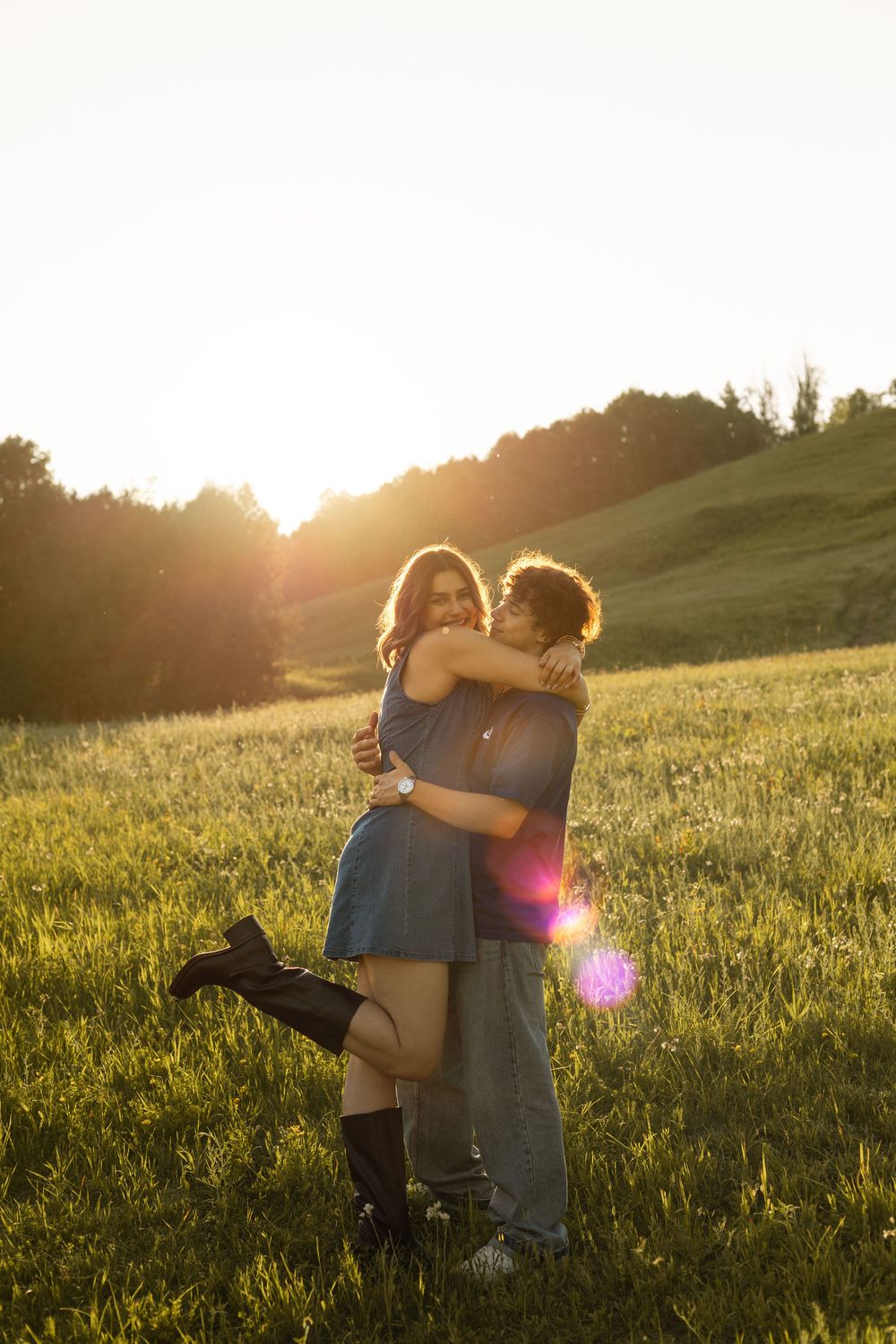 Couple embracing in a sunlit green field at golden hour, with warm lens flare and rolling hills in background.