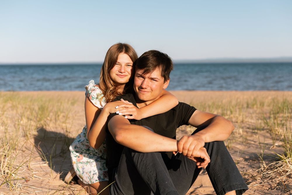 Couple sitting together on a sandy beach, woman hugging man from behind, smiling on a sunny day.