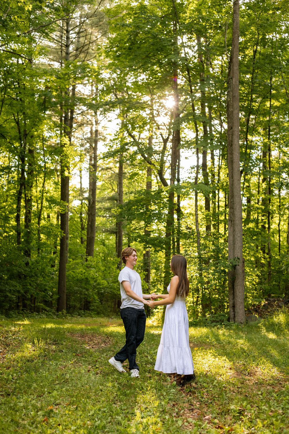 Couple holding hands in a sunlit forest clearing, woman in white dress and man in casual attire among tall green trees.