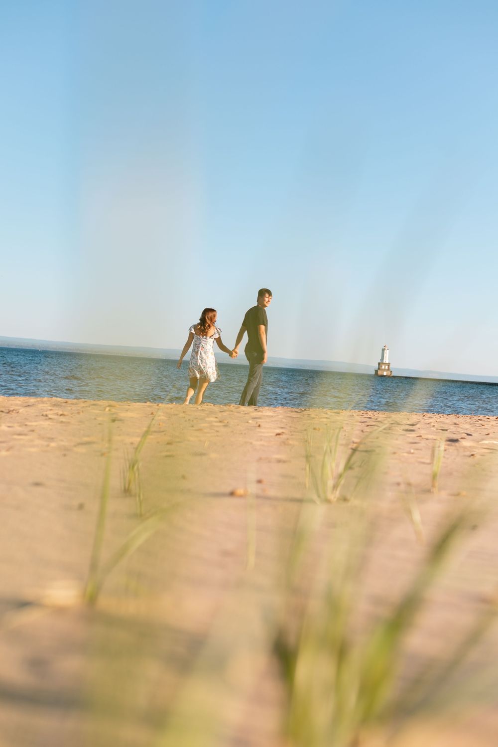 Couple holding hands walking on sandy beach with blue ocean and lighthouse in background on sunny day.