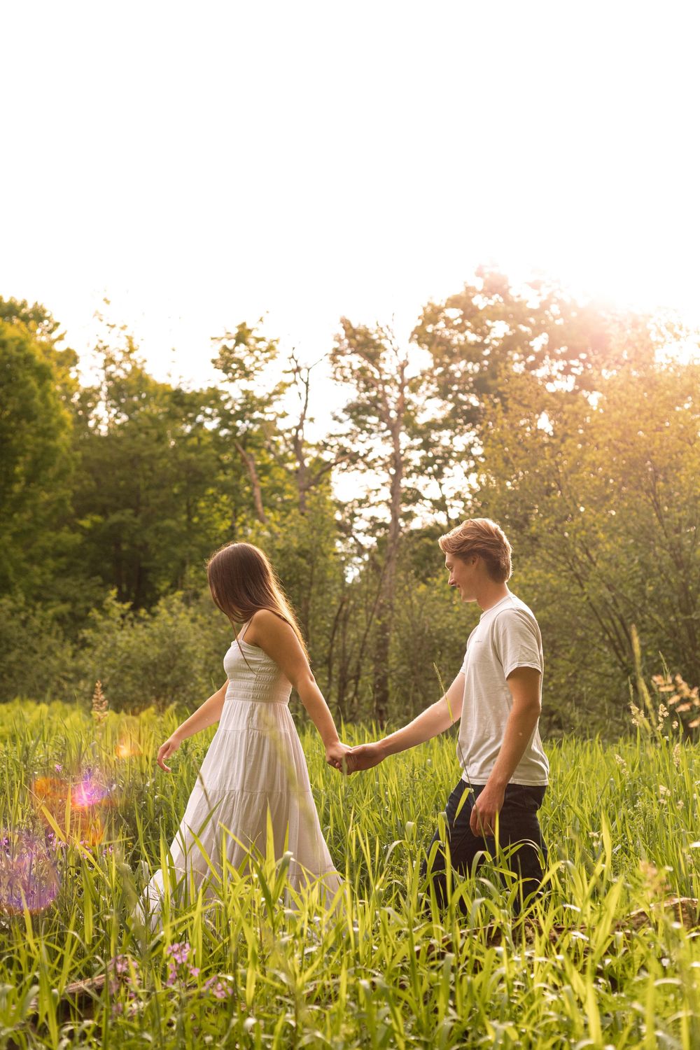 A couple holds hands walking through a lush green meadow with wildflowers at golden sunset.
