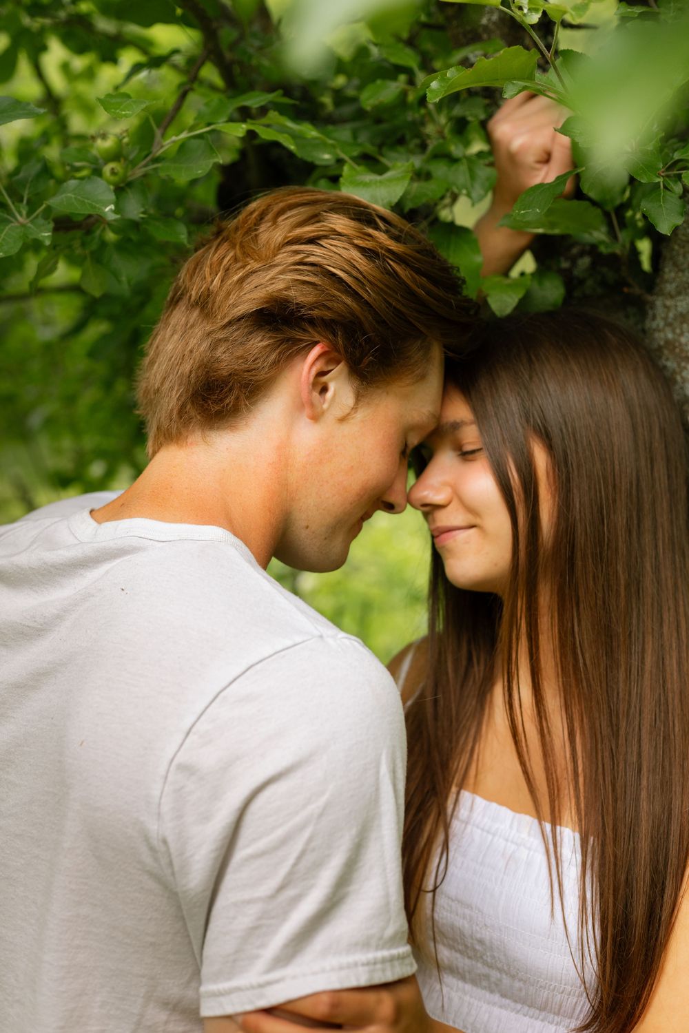 Young romantic couple touching foreheads tenderly under green tree leaves in a loving outdoor embrace.