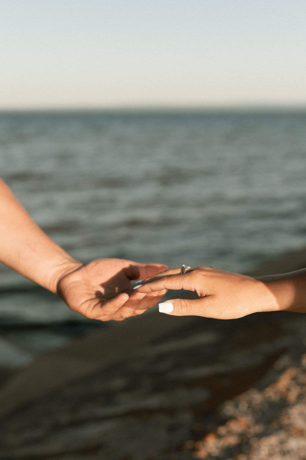 Two hands gently touching near the ocean shore, one adorned with a diamond engagement ring, symbolizing love and commitment.