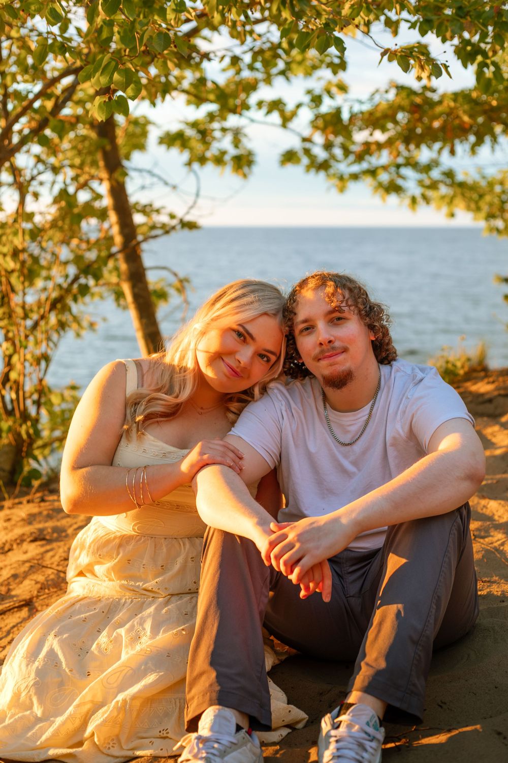 A young couple sitting together under a tree by the water during golden hour sunset, smiling warmly at the camera.