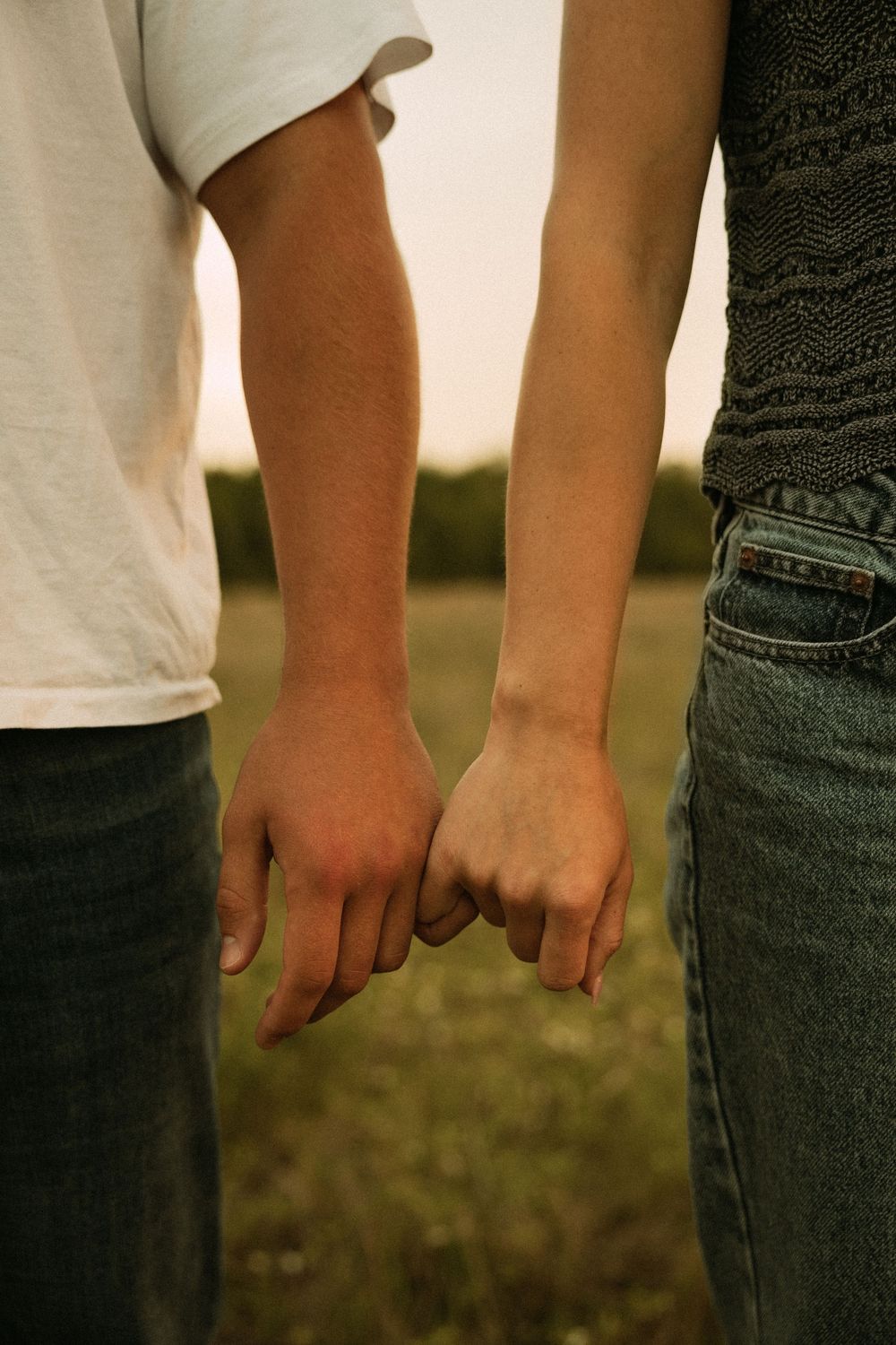 Two people holding pinkies in a field at sunset, wearing jeans and casual tops in a romantic outdoor setting.