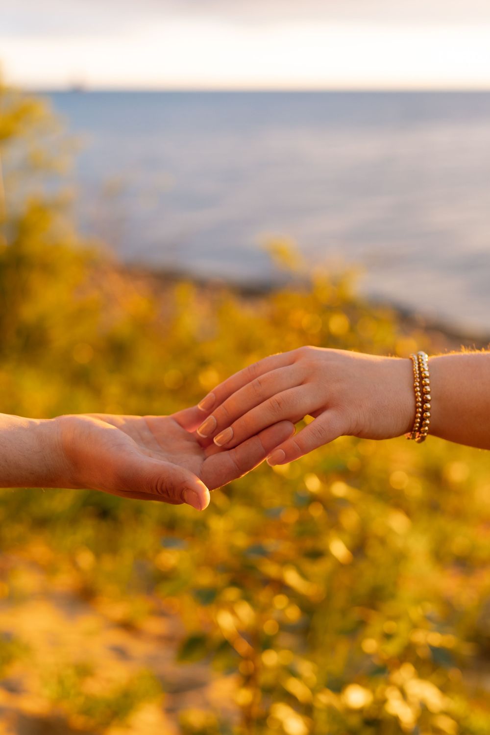 Two hands gently touching near a golden sunset shoreline, one adorned with bracelets, symbolizing love and connection.