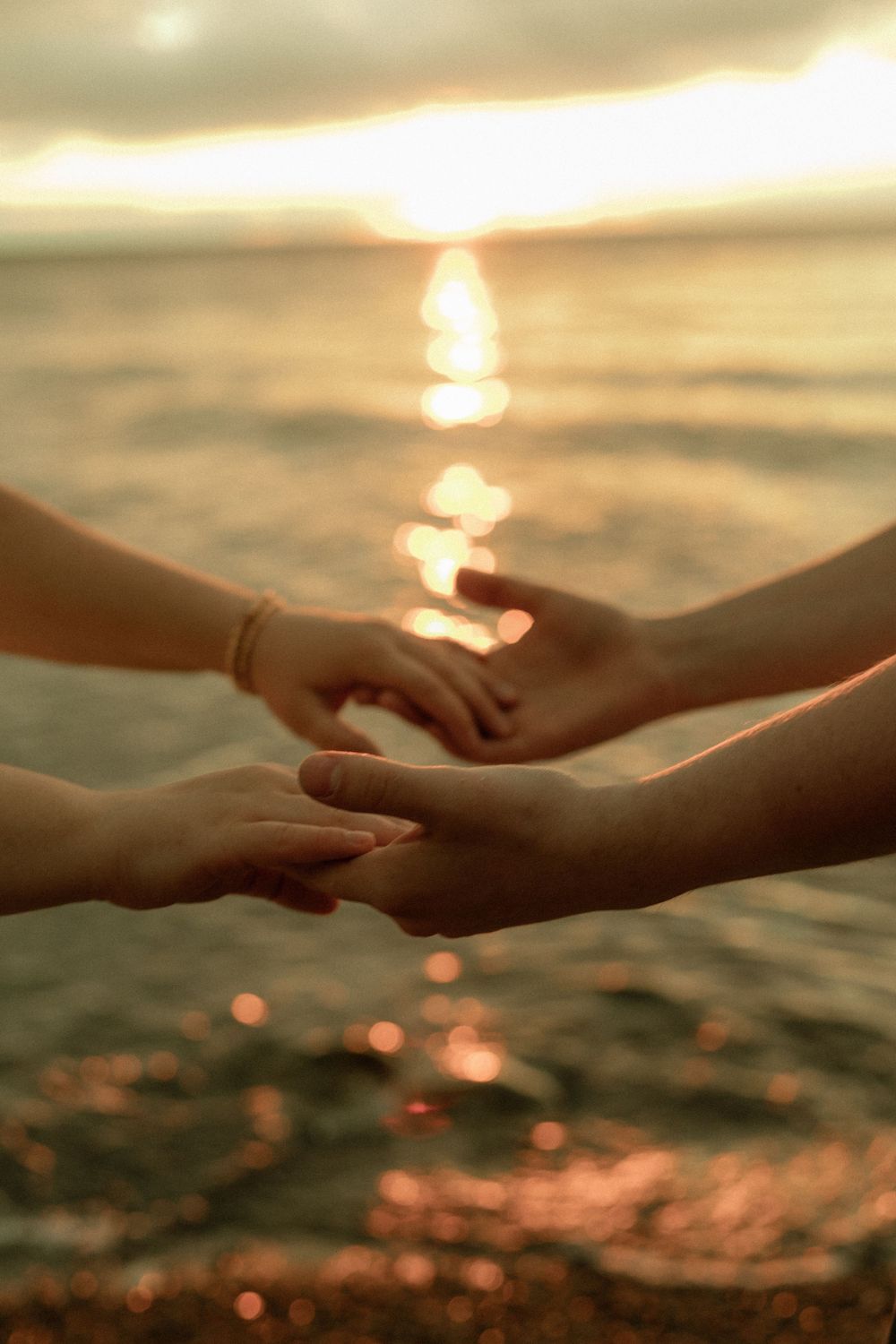 Two people gently holding hands at sunset over a shimmering ocean, with golden light reflecting on the water.