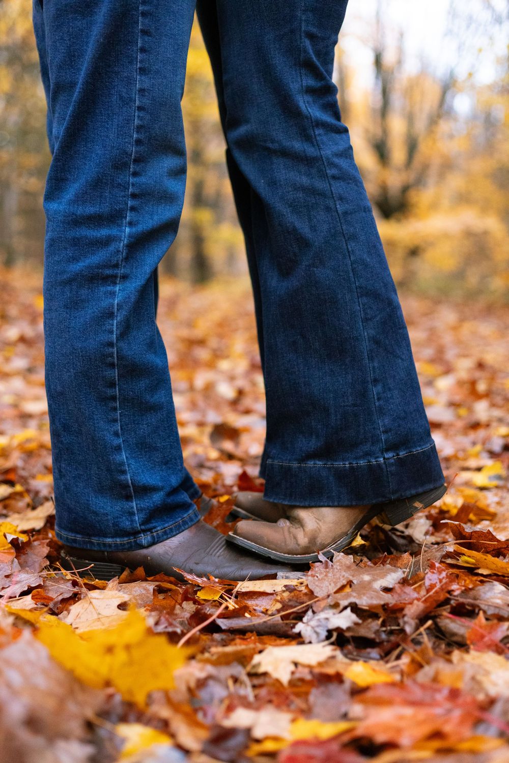 Feet in jeans and brown shoes walking through colorful fallen autumn leaves.