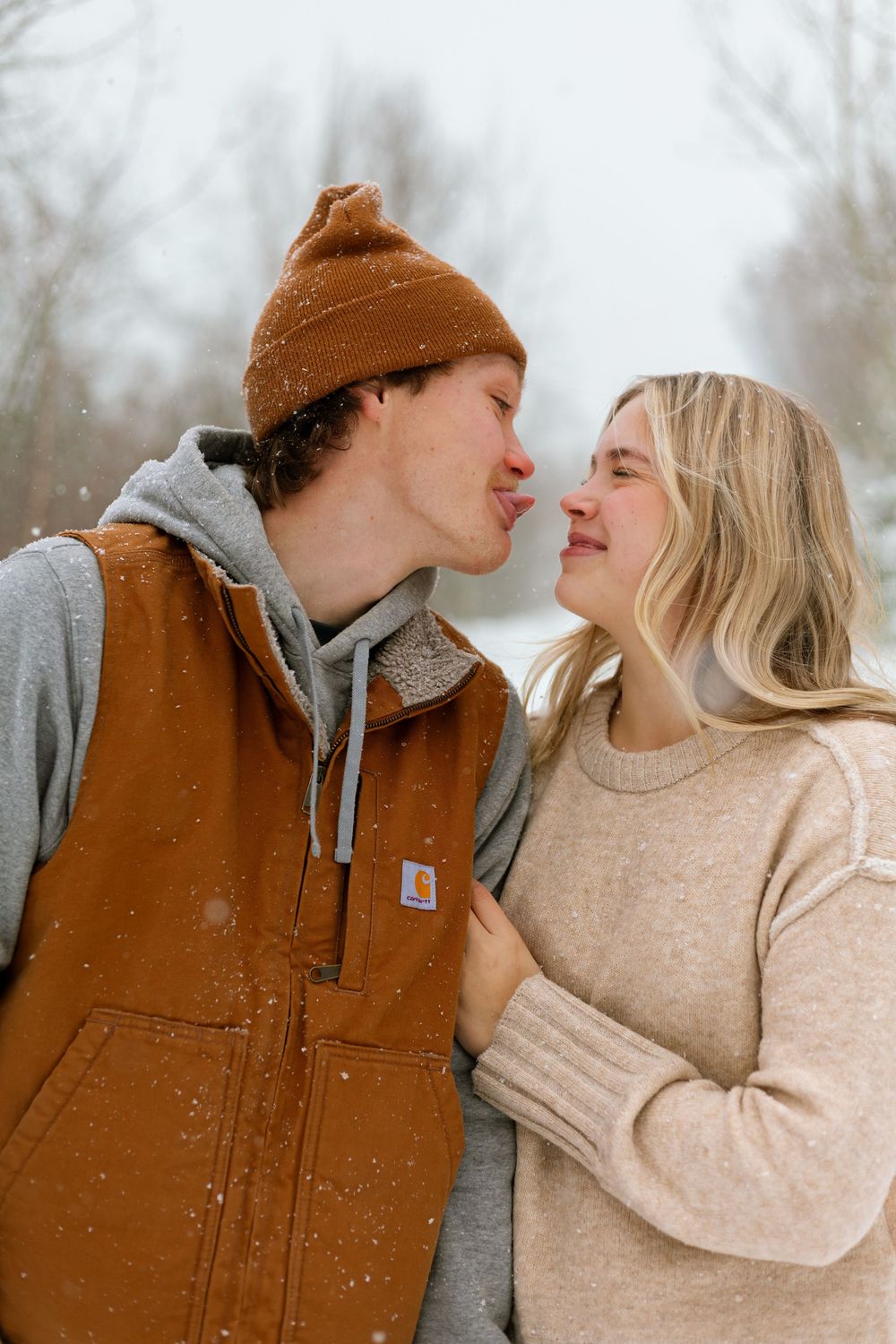 A couple shares intimate winter moments in a snowy scene wearing cozy knit sweaters and brown carhartt vest.