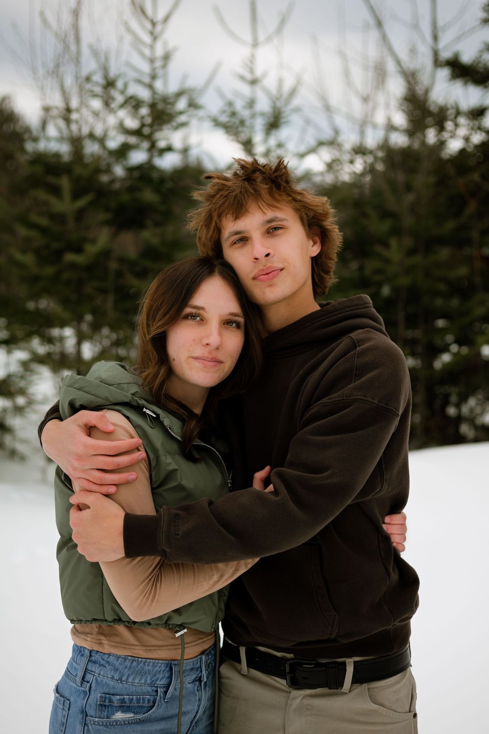 A couple embraces in front of snow-covered evergreen trees, wearing casual winter clothing.