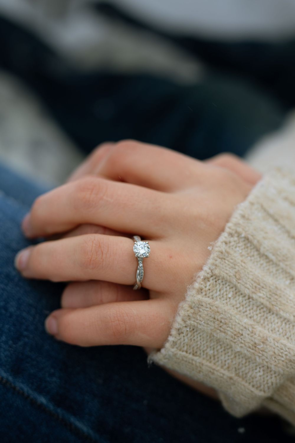 Close-up of a diamond solitaire engagement ring on a hand wearing a cream knit sweater against dark denim background.
