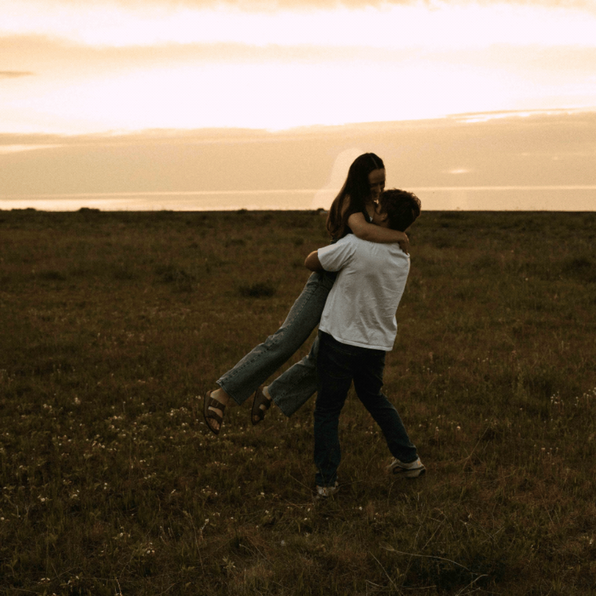 Couple embracing and dancing in a golden sunset field, romantic outdoor engagement photo.