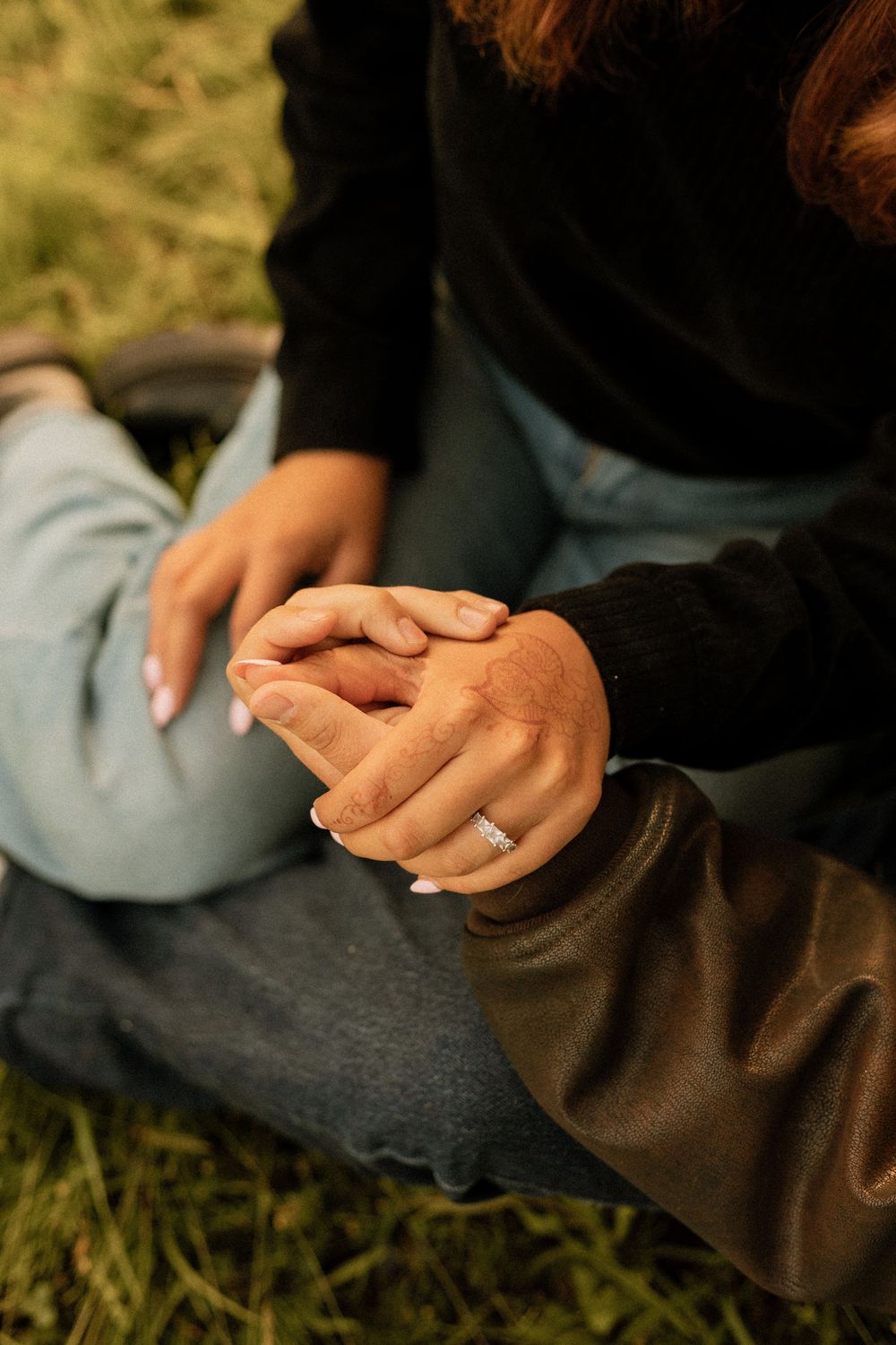 Close-up of couple holding hands on grass, woman wearing a diamond engagement ring, wearing jeans and dark sweaters.