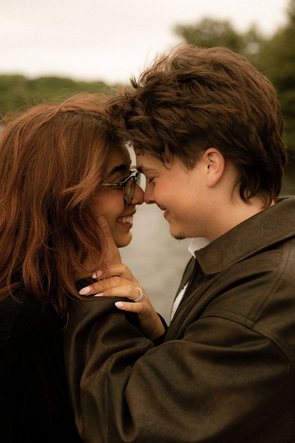A couple sharing a romantic moment, touching foreheads and smiling outdoors, woman wearing glasses and a ring.