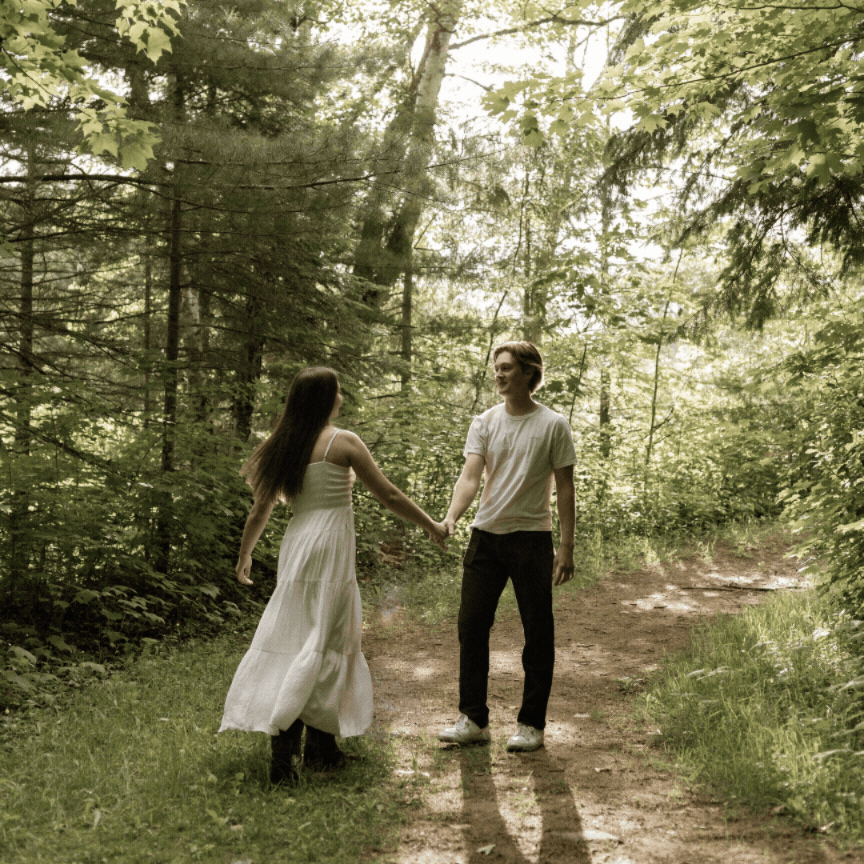 Couple holding hands on a forest path, woman in white dress, man in casual wear, surrounded by lush green trees.
