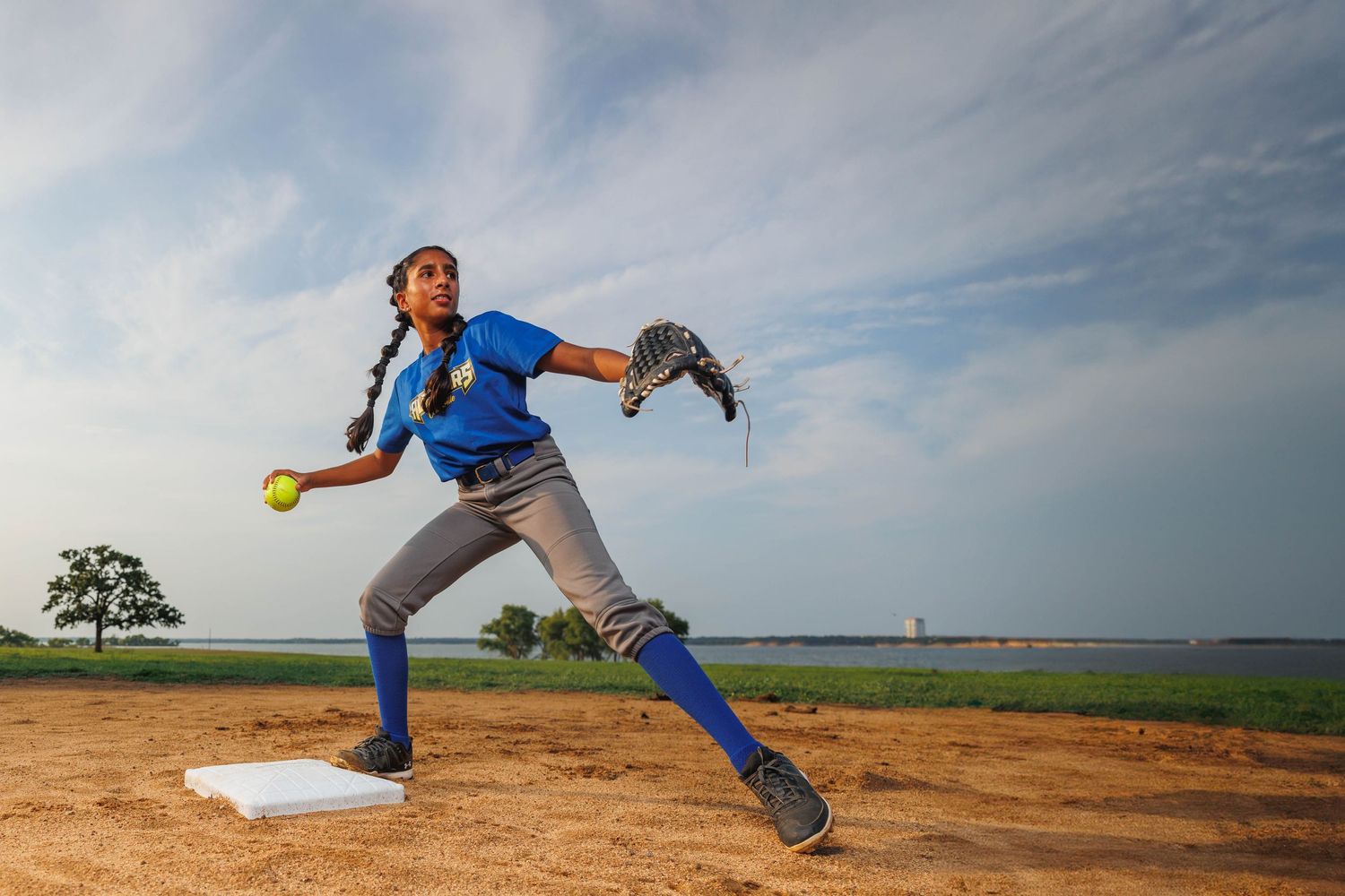 Softball pitcher in blue uniform and gray pants performs dynamic delivery motion on dirt mound against cloudy sky.
