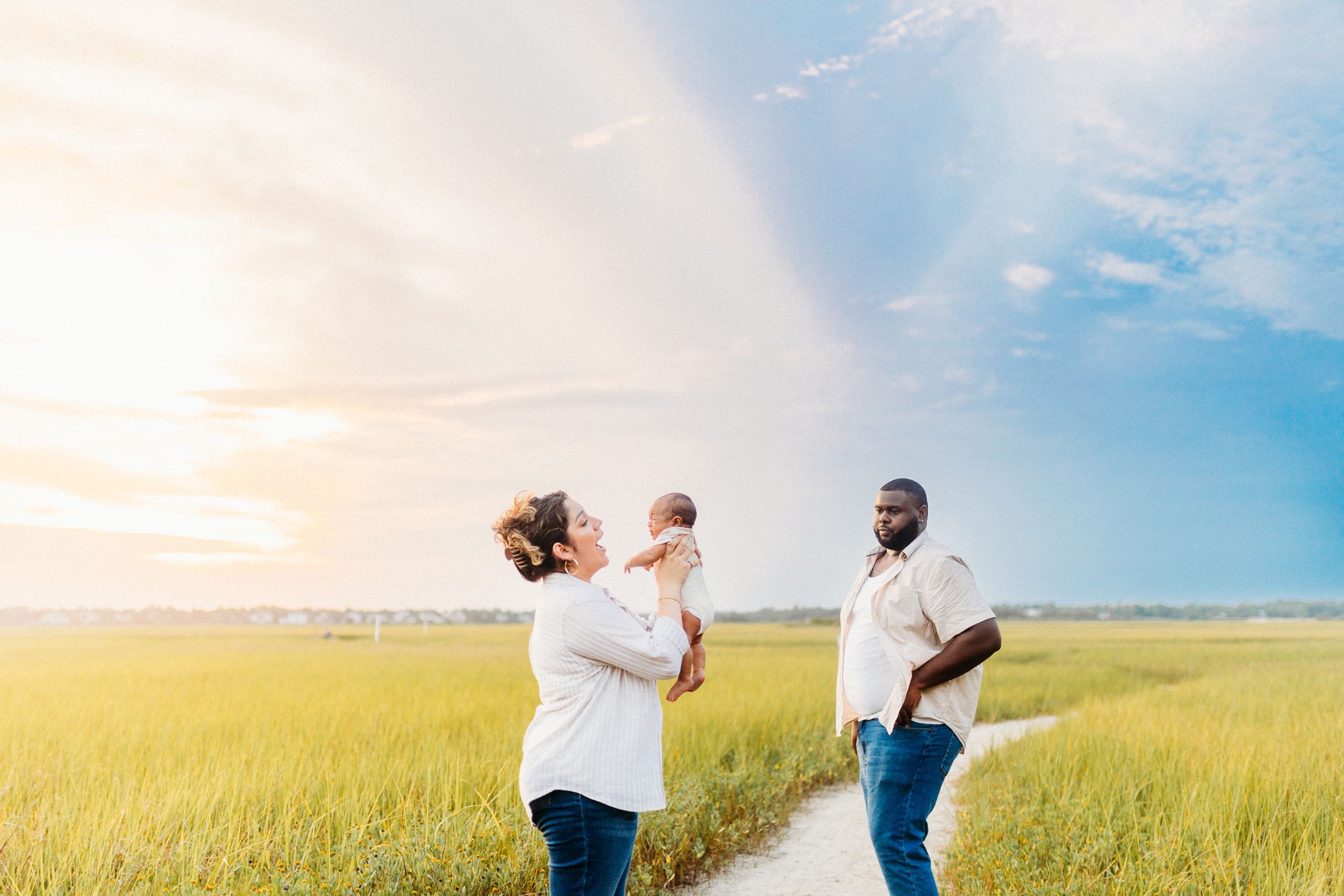 a mother holds her newborn baby boy in the air in the marsh at sunset, while dad smiles at them