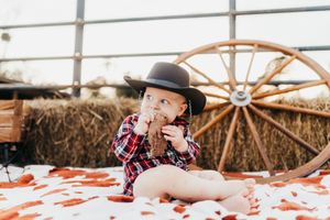 Baby wearing a black cowboy hat and plaid shirt sits on blanket with hay bales and wagon wheel in background.