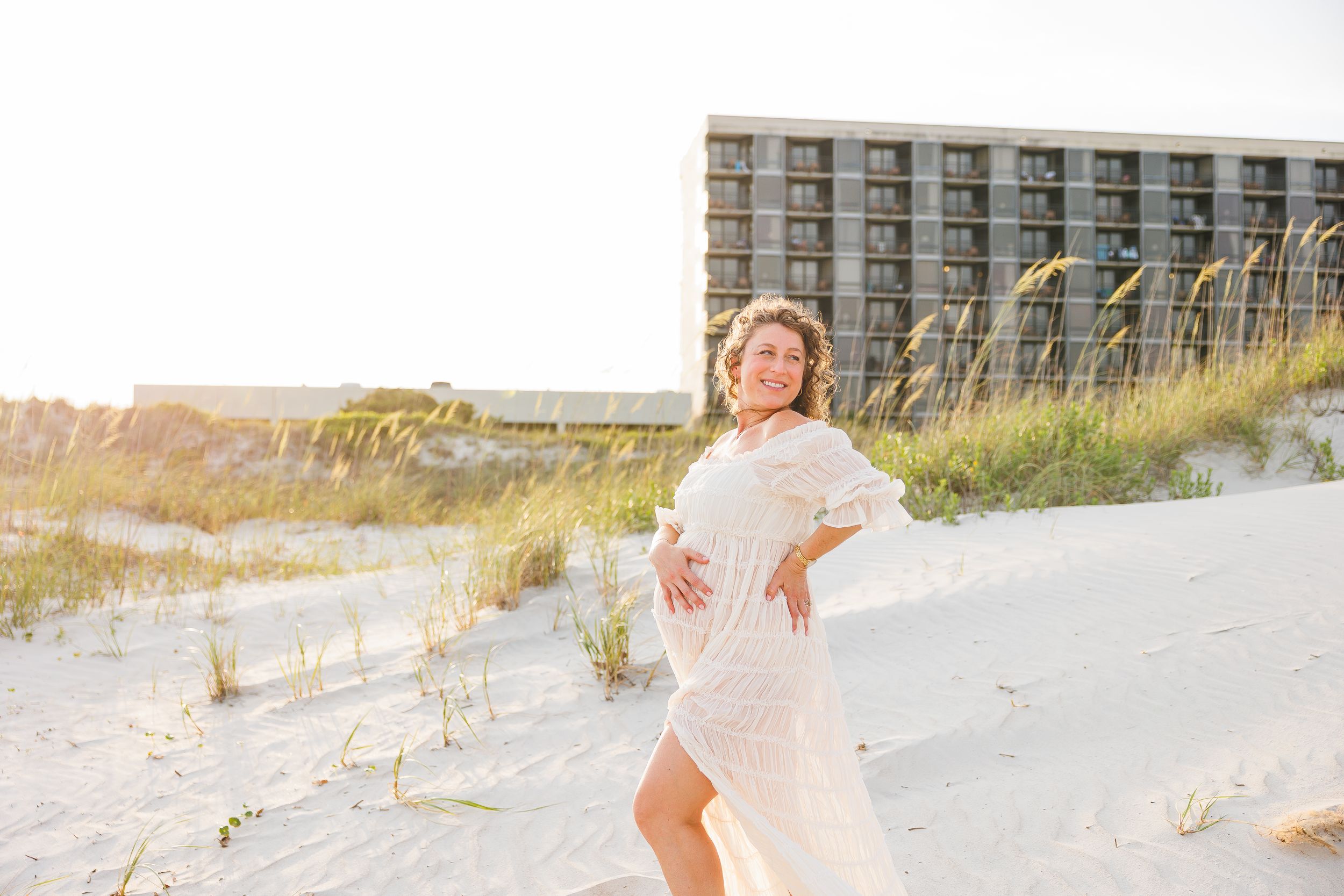 Pregnant woman in white lace dress posing on sandy beach dunes with sea oats and hotel in background.