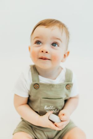 A baby wearing olive green overalls over a white shirt looks upward with bright eyes against a light background.