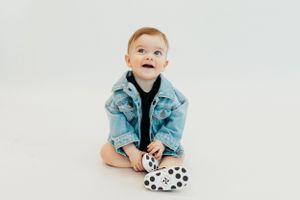 A small child wearing a denim jacket sits on a white background playing with a polka dot shoe.