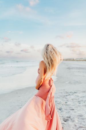 A person in a coral dress stands on a beach with flowing blonde hair during a dreamy sunset.