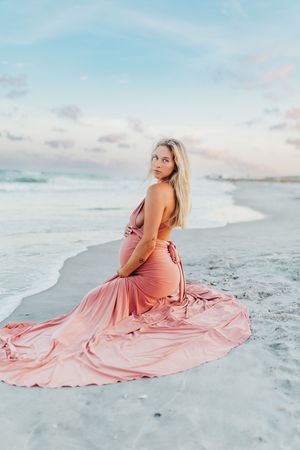 A person in a flowing pink dress poses on a white sandy beach at sunset with waves in the background.