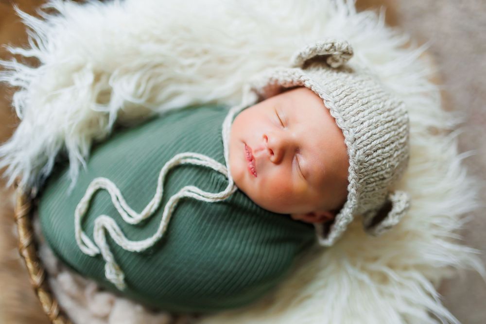 Newborn baby wearing sage green swaddle and knit bonnet lies on soft white fur blanket.