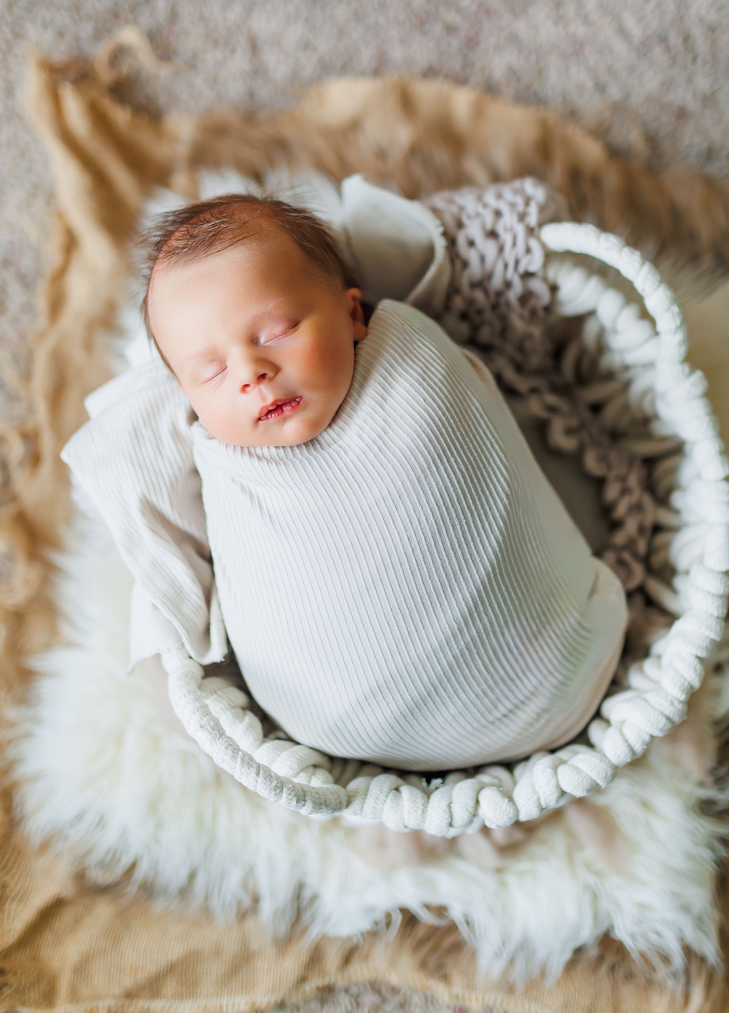 Sleeping newborn wrapped in white knit blanket rests peacefully in cozy furry basket on beige background.