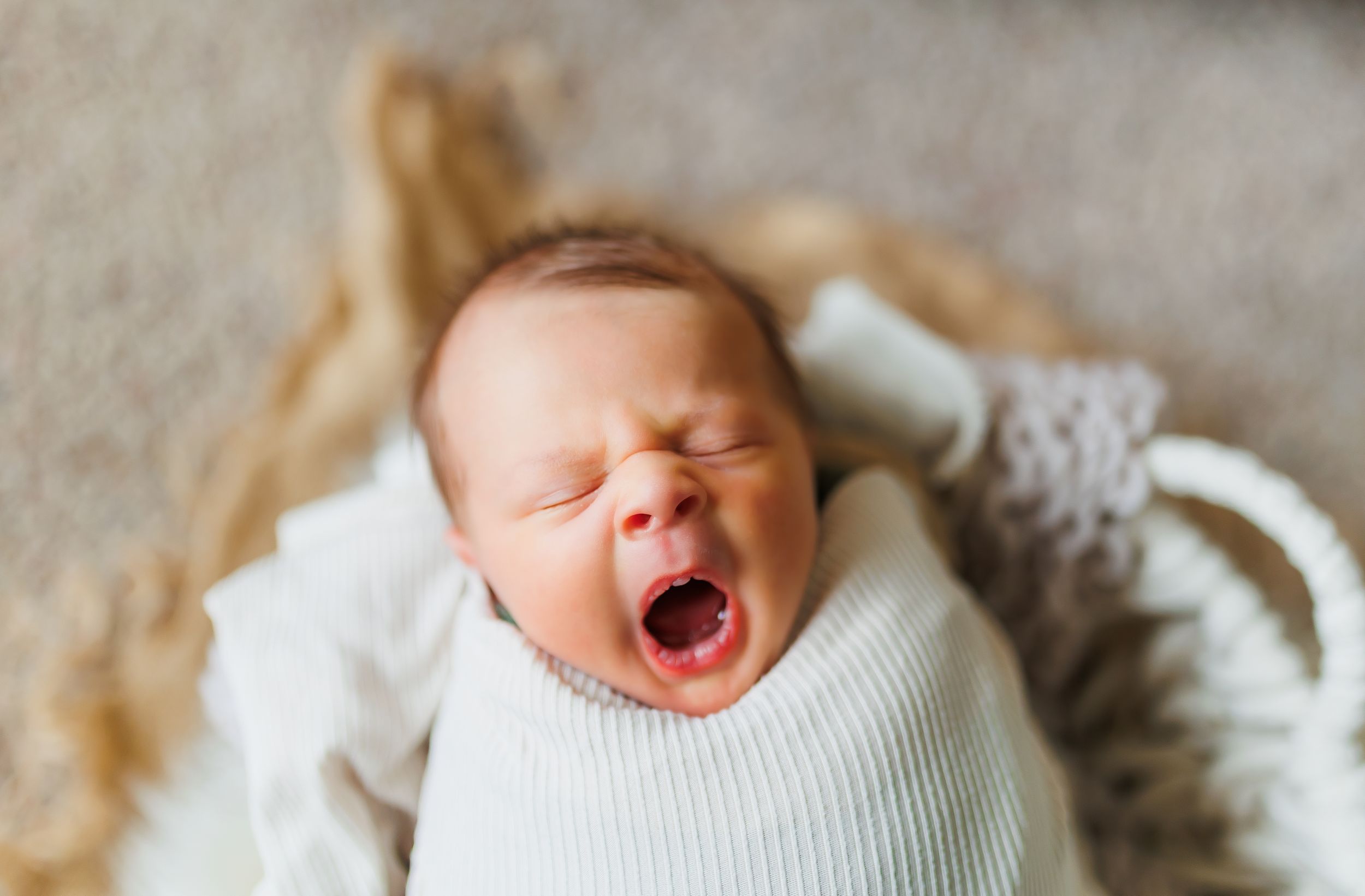 A tiny baby yawns while wearing a white sweater and lying on a soft textured blanket.