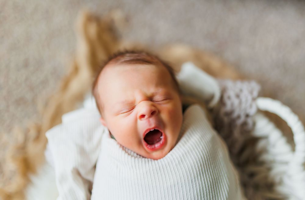 A tiny baby yawns while wearing a white sweater and lying on a soft textured blanket.