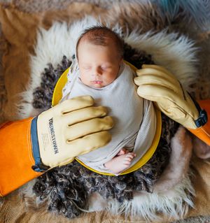 A sleeping baby swaddled in white lies on a fluffy sheepskin rug with yellow mittened hands visible.